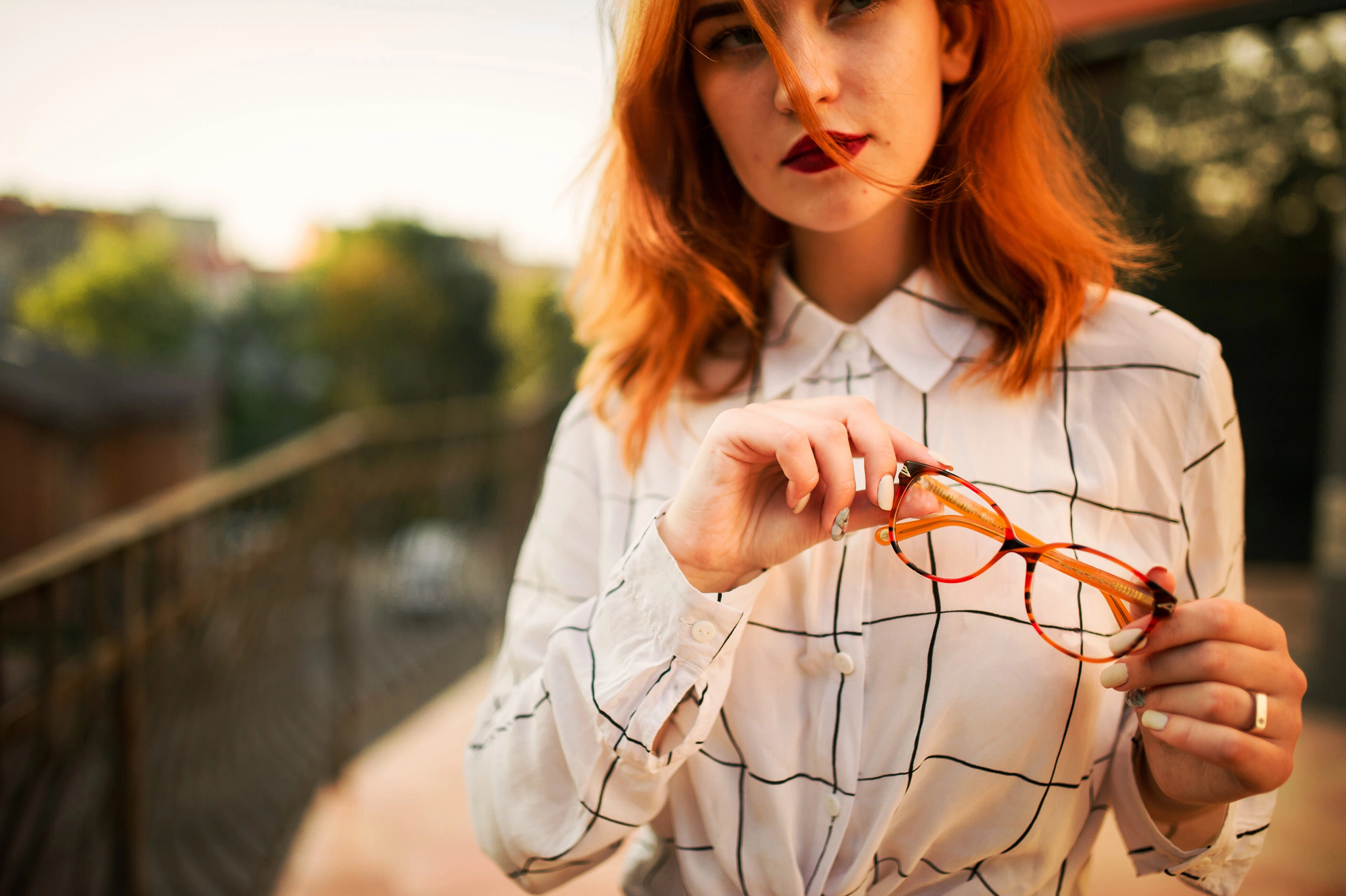 A woman with shoulder-length red hair stands outdoors, holding a pair of tortoiseshell glasses in her hand. She wears a white blouse with a black grid pattern. The background is blurred, featuring trees and buildings under a warm, golden light.