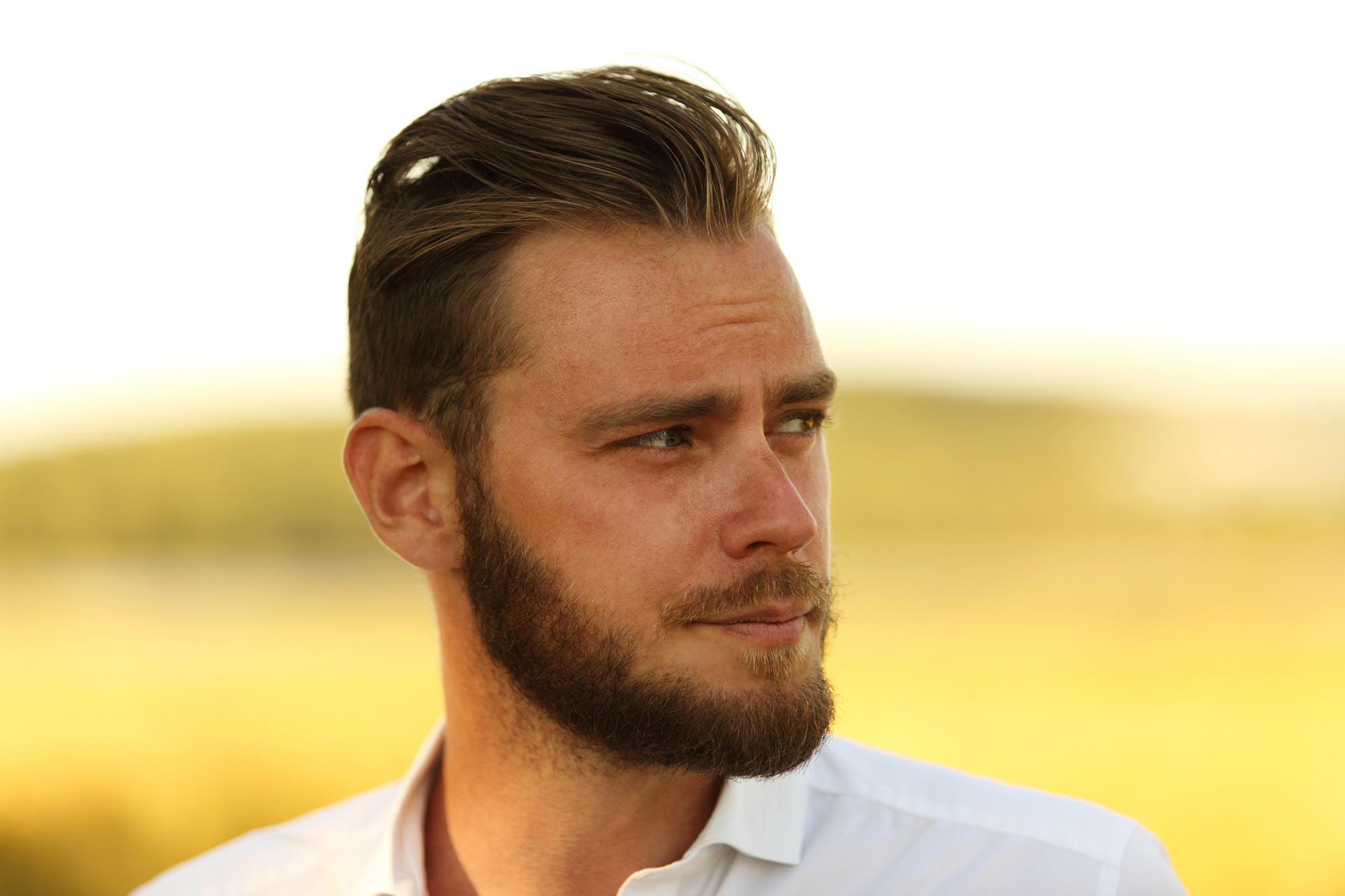 A man with short, styled hair and a beard gazes to his left. He is wearing a white shirt and standing outdoors with a blurred background of greenery and soft sunlight.