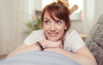 A young woman with red hair smiles and looks into the distance while leaning her chin on her folded arms. She is wearing a white shirt and has a bracelet on her wrist. The background is softly blurred, suggesting a cozy indoor setting with natural light.