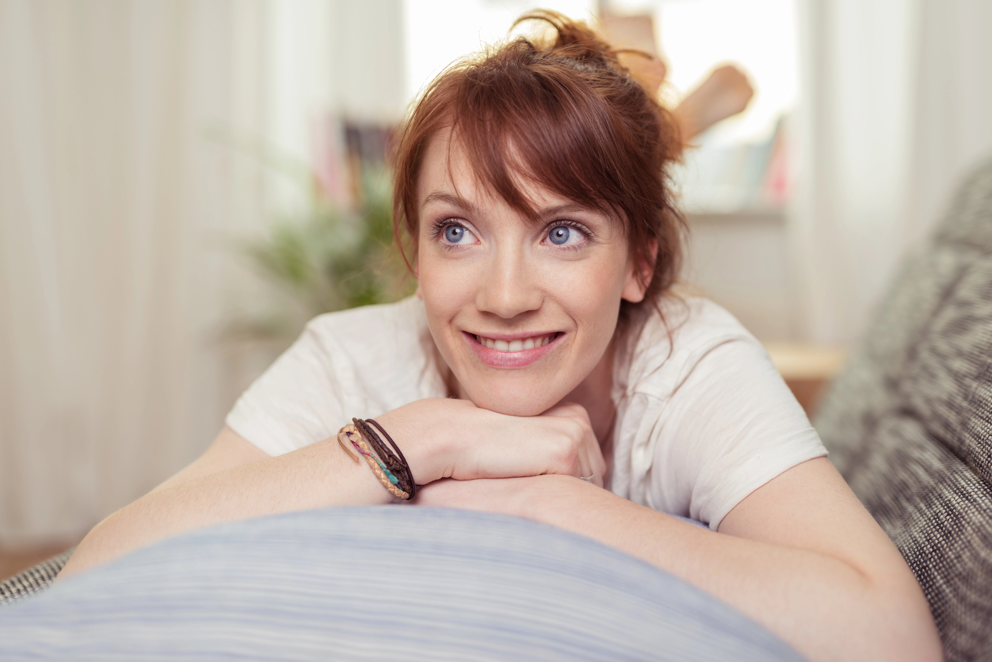 A young woman with red hair smiles and looks into the distance while leaning her chin on her folded arms. She is wearing a white shirt and has a bracelet on her wrist. The background is softly blurred, suggesting a cozy indoor setting with natural light.