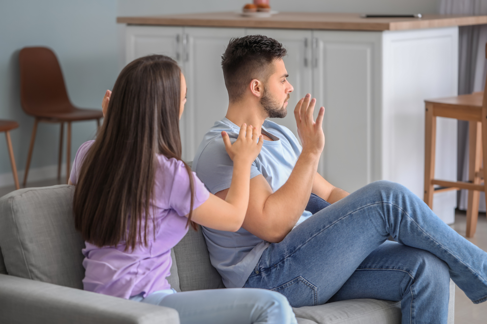 A woman with long brown hair sits on a couch, reaching out with an open hand towards a man with short dark hair. The man, sitting beside her, looks away with a serious expression, holding his hand up as if dismissing or stopping her. They appear to be in a living room.