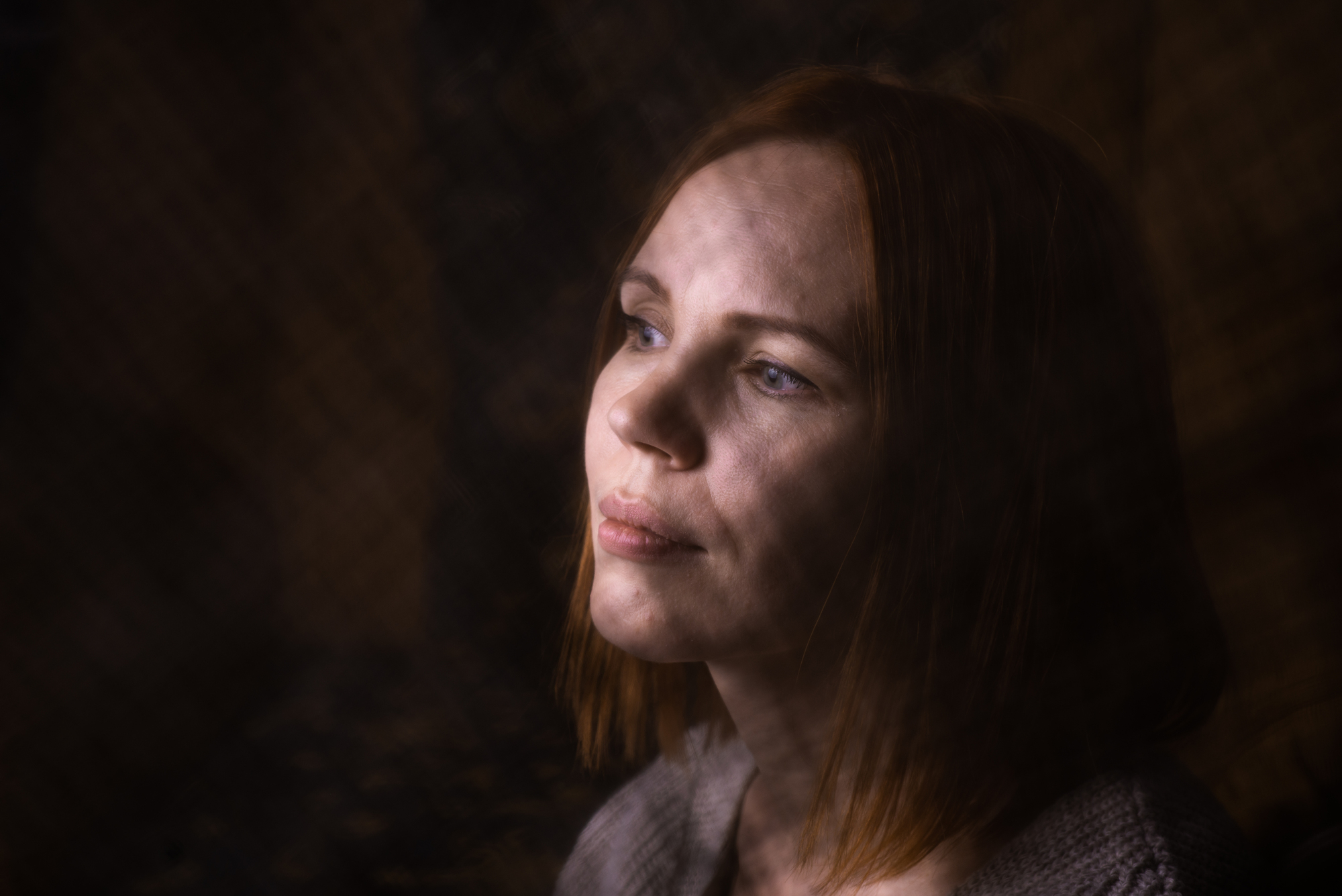 A woman with shoulder-length auburn hair gazes pensively into the distance in a dimly lit room. The soft lighting creates shadows on her face, highlighting her thoughtful expression and serene demeanor.