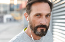 A man with short brown hair and a beard, wearing a grey shirt, is looking directly at the camera while wearing white earphones. The background is blurred, suggesting an outdoor setting with urban elements. The lighting is soft, coming from the left side.