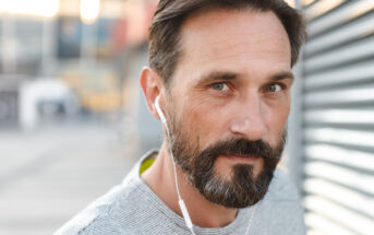 A man with short brown hair and a beard, wearing a grey shirt, is looking directly at the camera while wearing white earphones. The background is blurred, suggesting an outdoor setting with urban elements. The lighting is soft, coming from the left side.