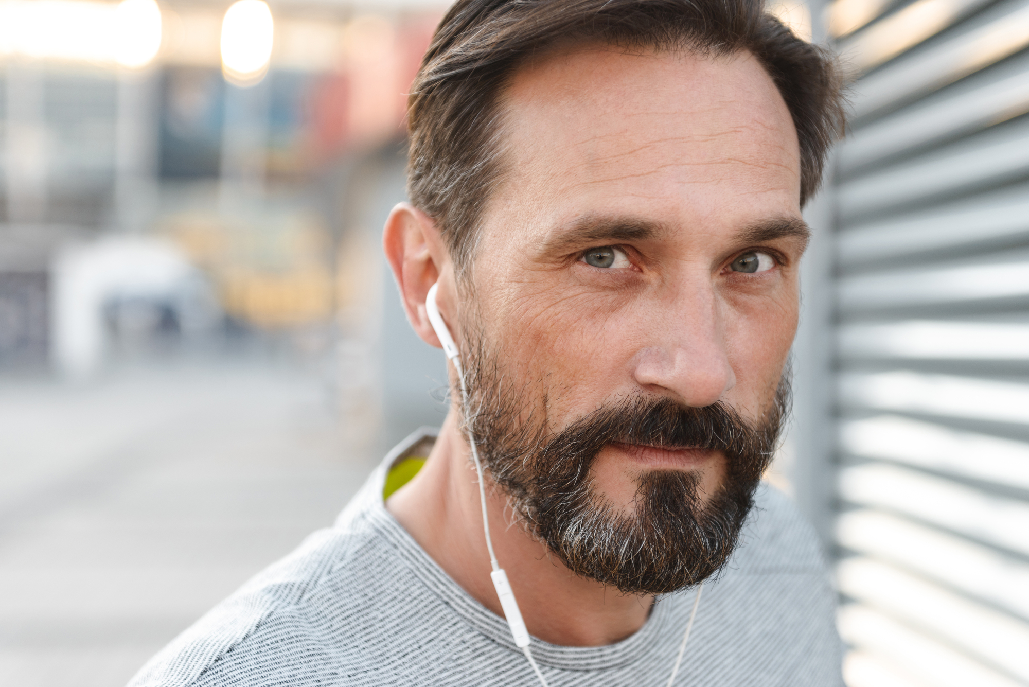 A man with short brown hair and a beard, wearing a grey shirt, is looking directly at the camera while wearing white earphones. The background is blurred, suggesting an outdoor setting with urban elements. The lighting is soft, coming from the left side.