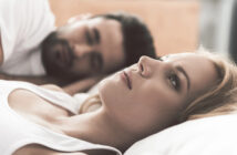 A woman with blonde hair and a serious expression lies on a bed, staring thoughtfully upward. Beside her, a man with dark hair and beard lies close, looking at her with a soft expression. Both are dressed in casual white tops in a relaxed bedroom setting.