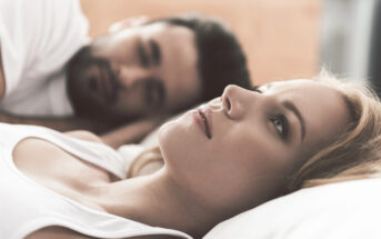 A woman with blonde hair and a serious expression lies on a bed, staring thoughtfully upward. Beside her, a man with dark hair and beard lies close, looking at her with a soft expression. Both are dressed in casual white tops in a relaxed bedroom setting.