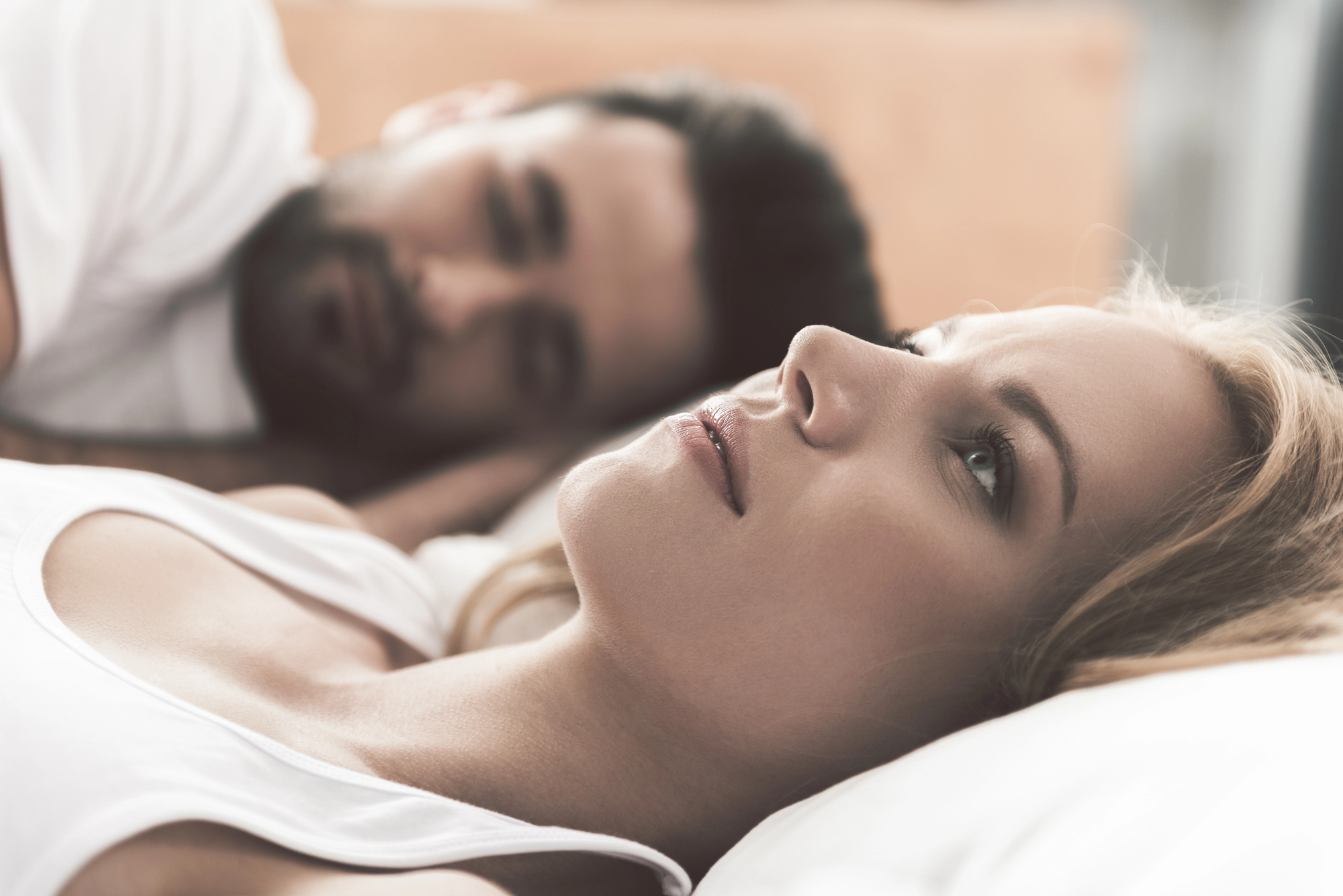 A woman with blonde hair and a serious expression lies on a bed, staring thoughtfully upward. Beside her, a man with dark hair and beard lies close, looking at her with a soft expression. Both are dressed in casual white tops in a relaxed bedroom setting.