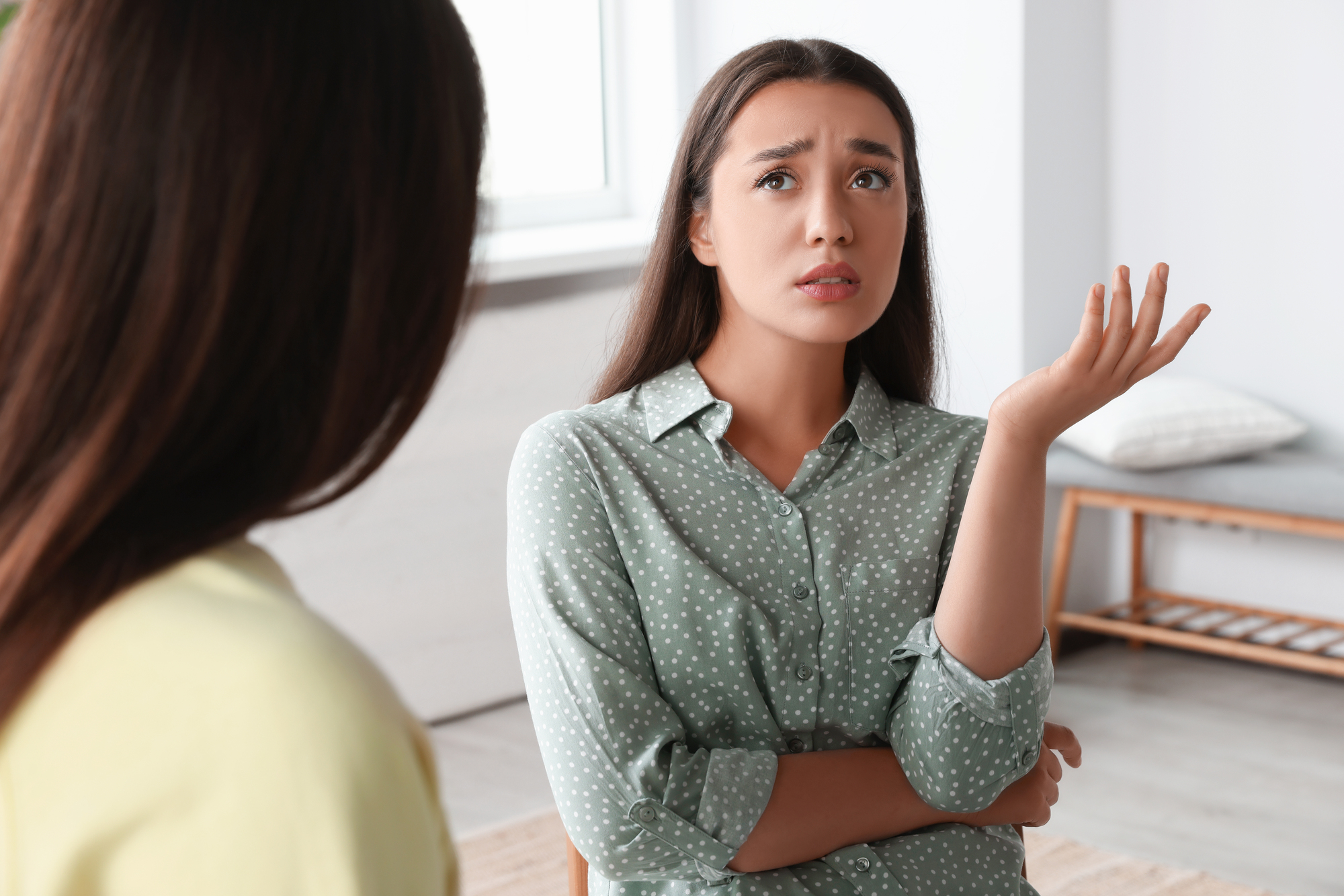 A woman with long dark hair, wearing a green polka dot shirt, looks concerned and raises one hand in a questioning gesture while talking to another person whose back is facing the camera. They are indoors with a bench and cushion in the background.