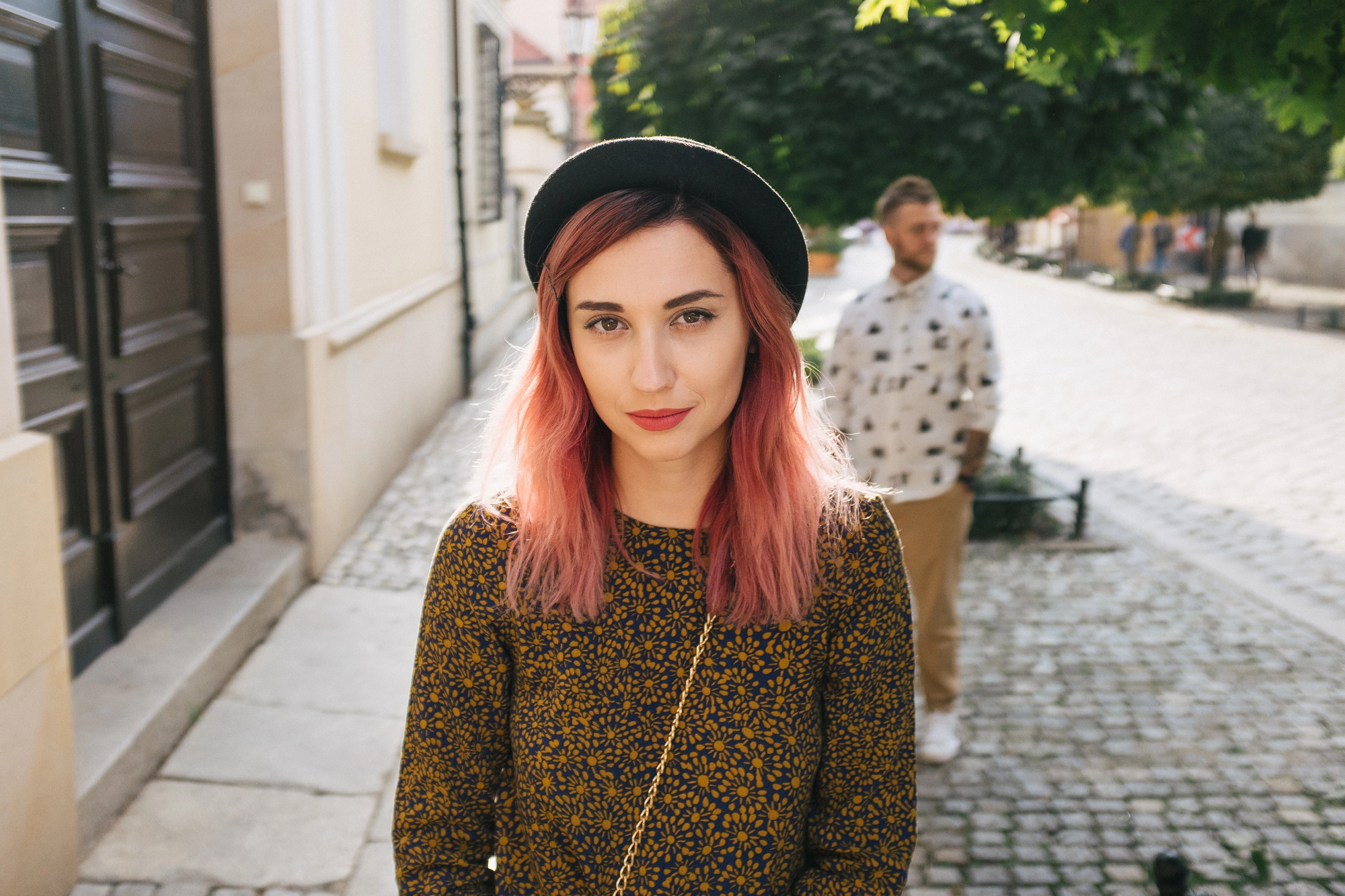 A woman with pink hair, wearing a black hat and a patterned mustard-colored top, stands on a cobblestone street. A man in a patterned shirt and beige pants stands in the background, slightly out of focus. Trees and buildings line the sunny street.