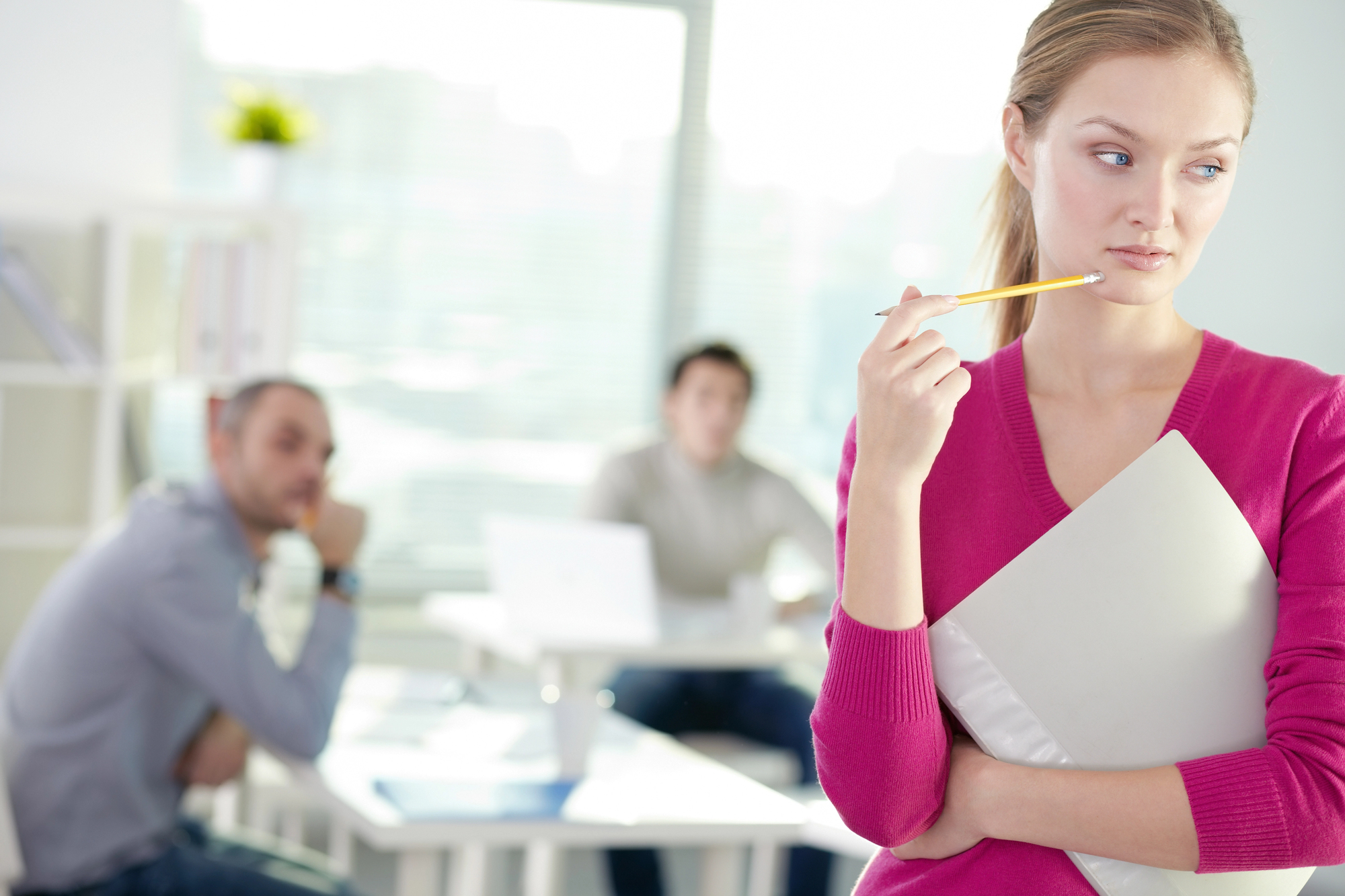 A woman in a pink sweater holds a folder and a pencil, looking pensive. Behind her, a man and woman sit at a desk, blurred in the background. The room appears to be an office with bright daylight streaming through the windows.