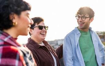 Three people are standing outside and smiling. The person on the left is wearing a red plaid shirt, the person in the middle is wearing sunglasses and a dark jacket, and the person on the right is wearing glasses, a light blue shirt, and a green top. The background is slightly blurred.