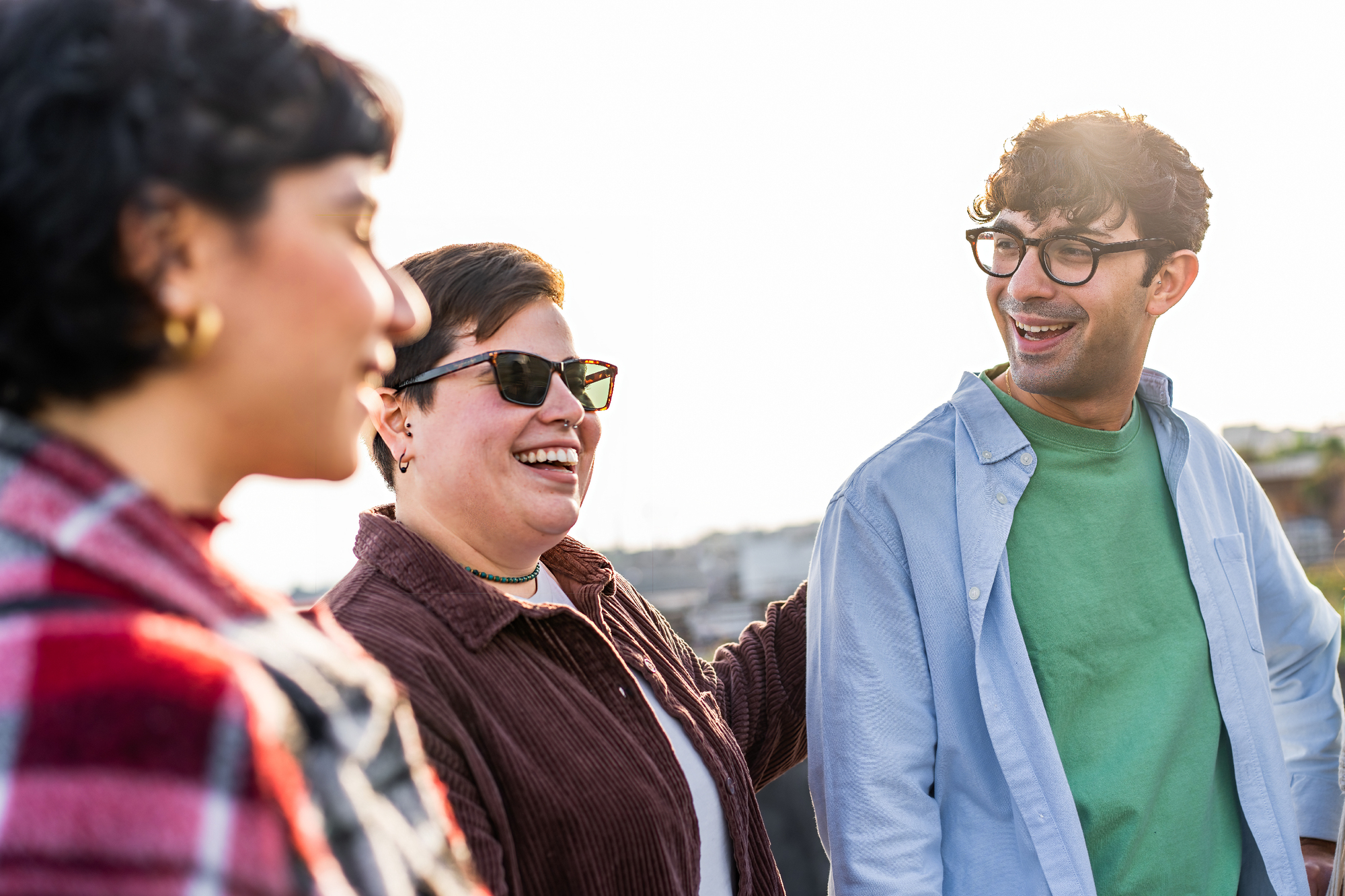 Three people are standing outside and smiling. The person on the left is wearing a red plaid shirt, the person in the middle is wearing sunglasses and a dark jacket, and the person on the right is wearing glasses, a light blue shirt, and a green top. The background is slightly blurred.