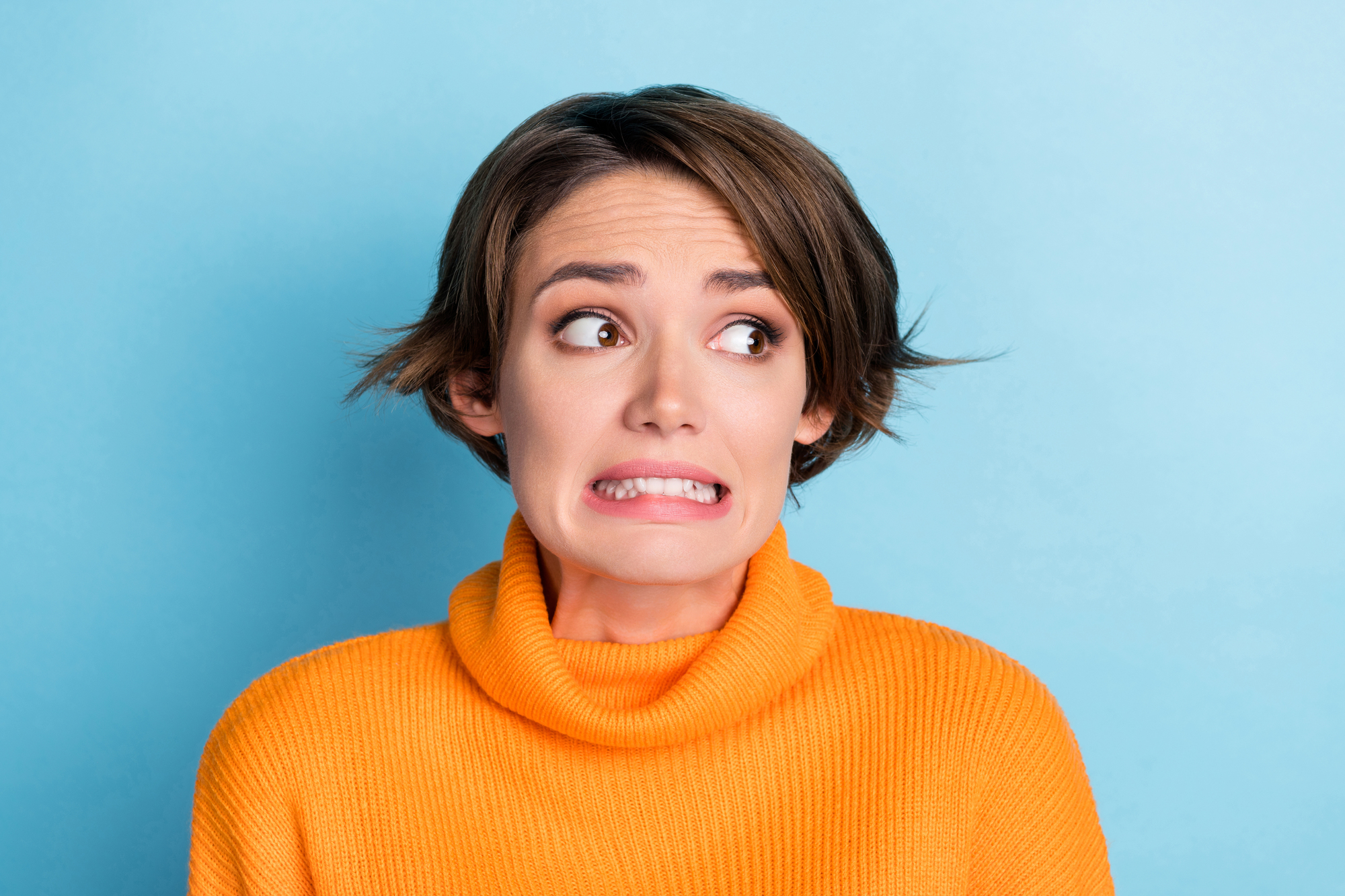 A person with short brown hair wearing an orange sweater is standing against a light blue background. They have a worried or nervous expression with wide eyes and a grimacing mouth.