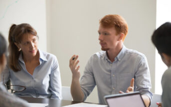 A man with red hair and a beard, dressed in a light blue shirt, gestures and speaks during a meeting. A woman with long brown hair, also wearing a light blue shirt, listens attentively. Other people are seated at the table with notepads and laptops visible.