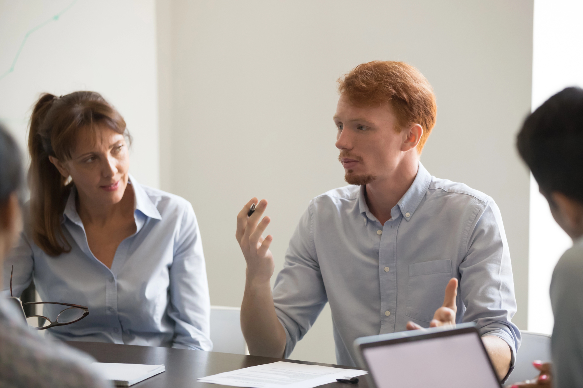 A man with red hair and a beard, dressed in a light blue shirt, gestures and speaks during a meeting. A woman with long brown hair, also wearing a light blue shirt, listens attentively. Other people are seated at the table with notepads and laptops visible.