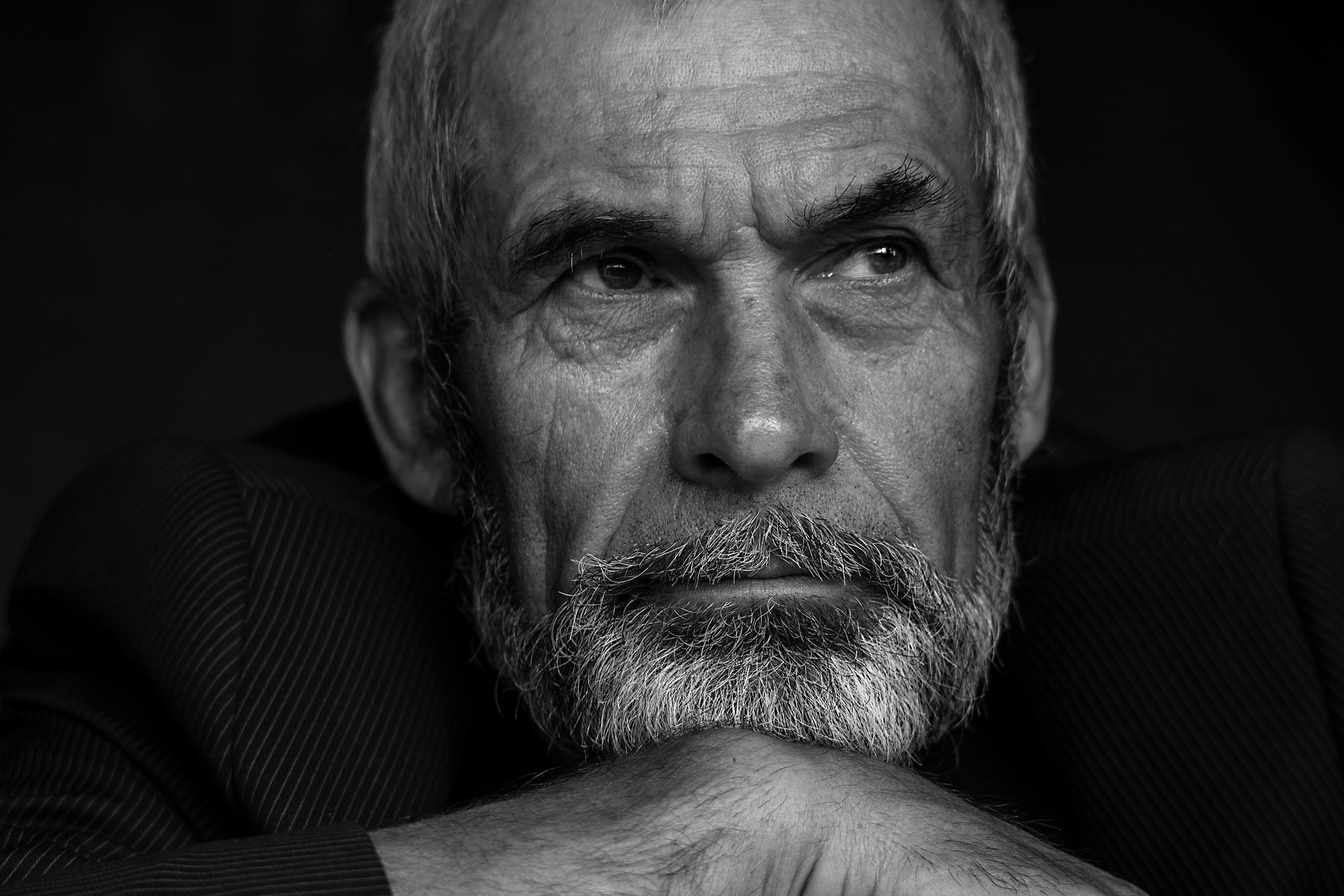 Black and white portrait of an elderly man with a thoughtful expression. He has a full grey beard and hair, and is resting his chin on his crossed arms. He wears a pinstriped suit jacket and looks slightly to the side, with a pensive gaze.