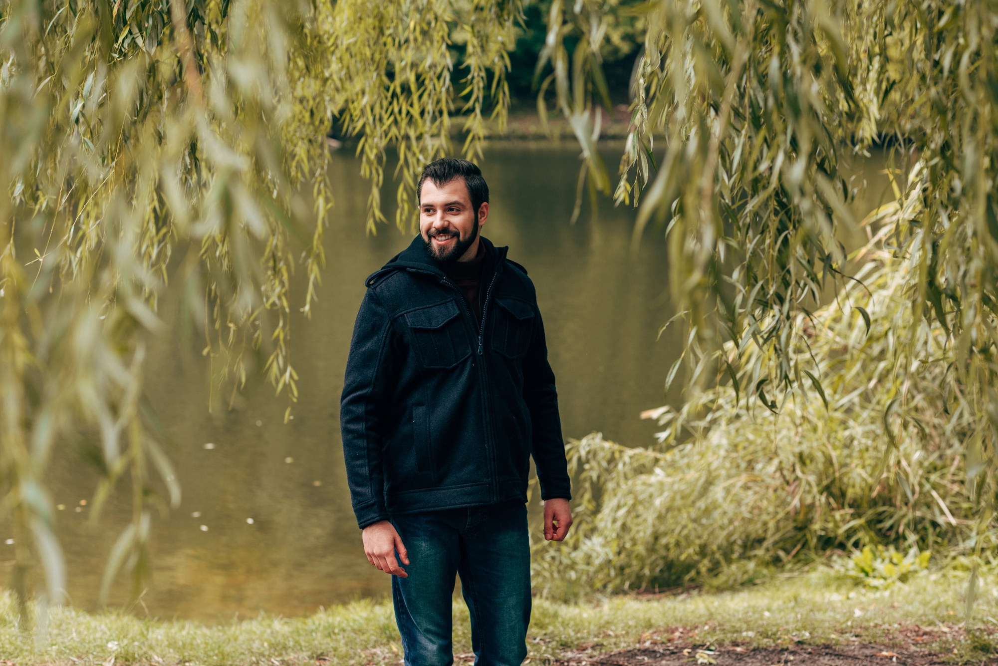 A man with a beard stands near a body of water framed by overhanging willow branches. He is wearing a dark jacket and jeans, smiling and looking off to the side. The background features lush greenery.