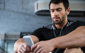 A man with short dark hair and a beard is sitting in a gym, wearing a black athletic shirt, and looking at his smartphone. He has earphones in his ears and appears focused or contemplative. The gym equipment and a large air conditioning unit are visible in the background.