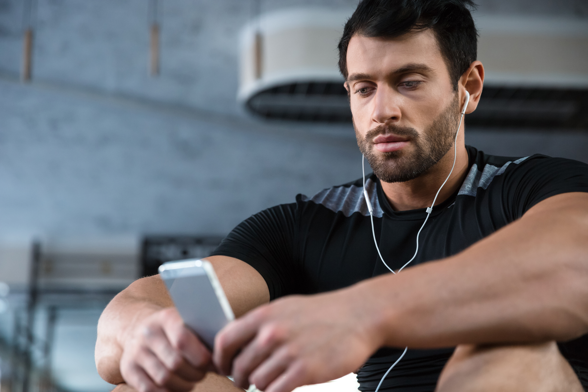 A man with short dark hair and a beard is sitting in a gym, wearing a black athletic shirt, and looking at his smartphone. He has earphones in his ears and appears focused or contemplative. The gym equipment and a large air conditioning unit are visible in the background.