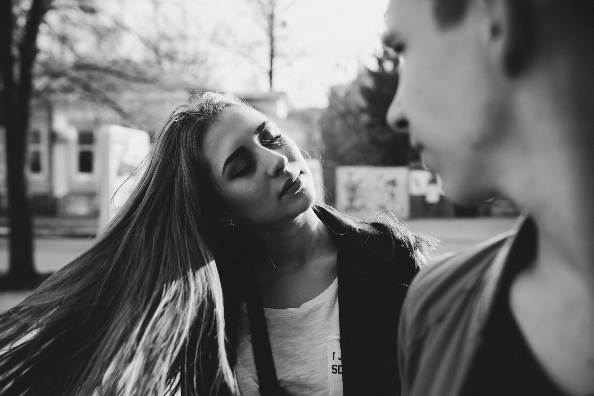 Black and white photo of two people outside. The person in the foreground is out of focus, while the second person, in focus, has long hair flowing as they tilt their head back and look away. Trees and houses are blurred in the background.