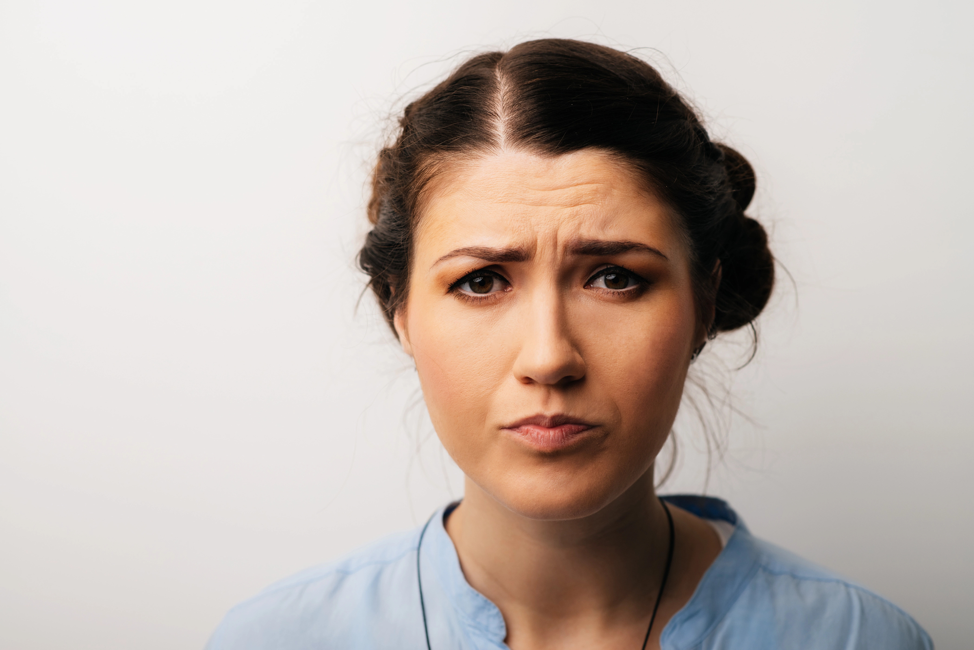 A woman with brown hair styled in two braided buns stands against a plain white background. She has a concerned or perplexed expression on her face with furrowed eyebrows. She is wearing a light blue top with a V-neckline.