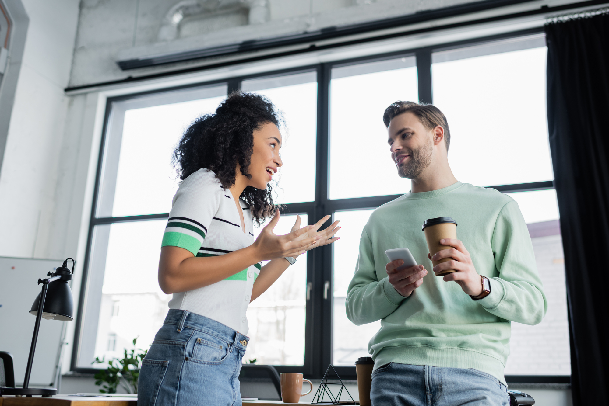 A woman and a man are having a conversation in an office. The woman, animated and gesturing with her hands, is wearing a white shirt and jeans. The man, holding a smartphone and a coffee cup, is wearing a light green sweater and blue jeans. Large windows are visible behind them.