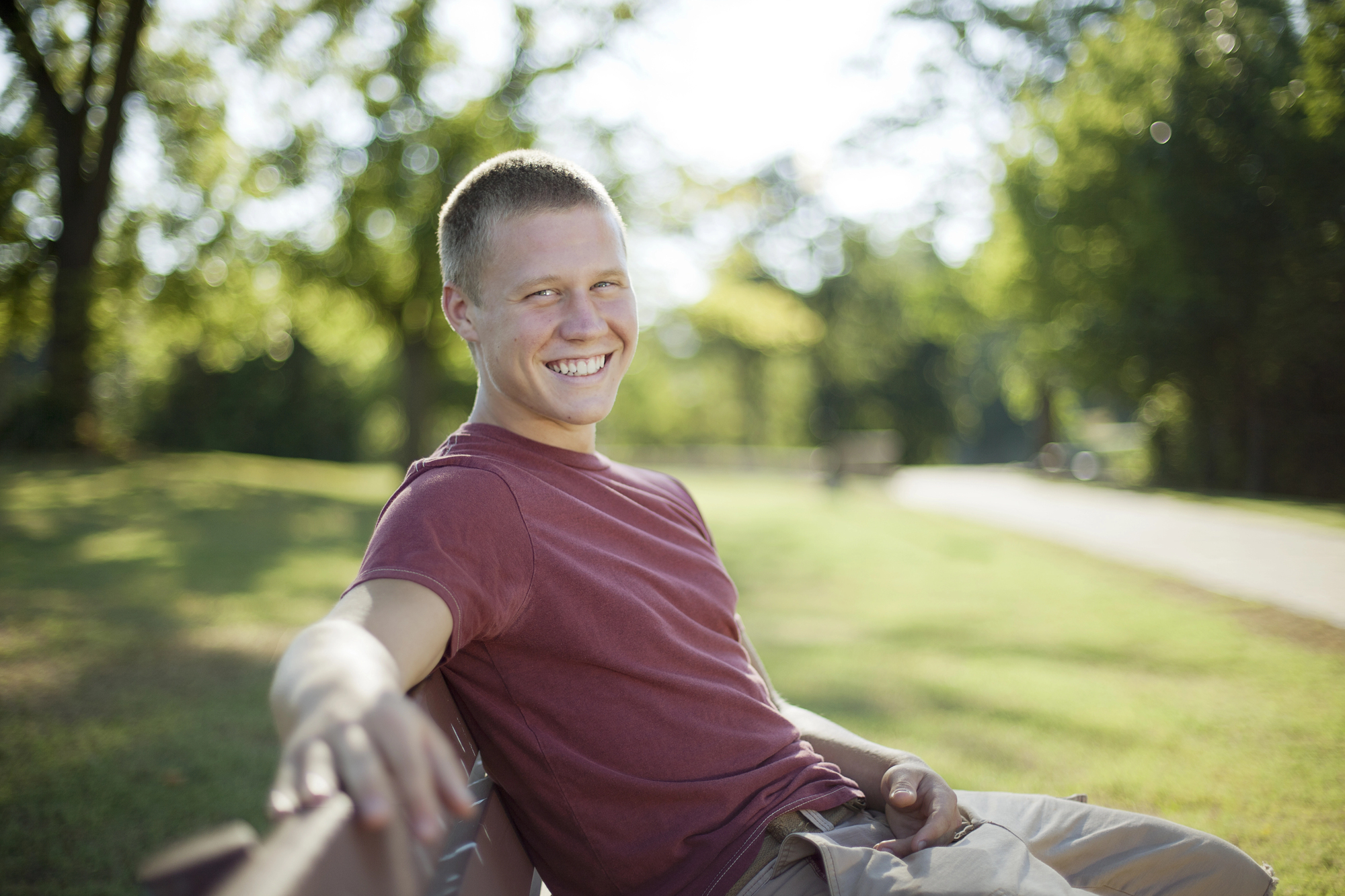 A young man with short hair and wearing a red shirt is sitting on a park bench, smiling at the camera. The background features green trees and grass, indicating a sunny day in the park.