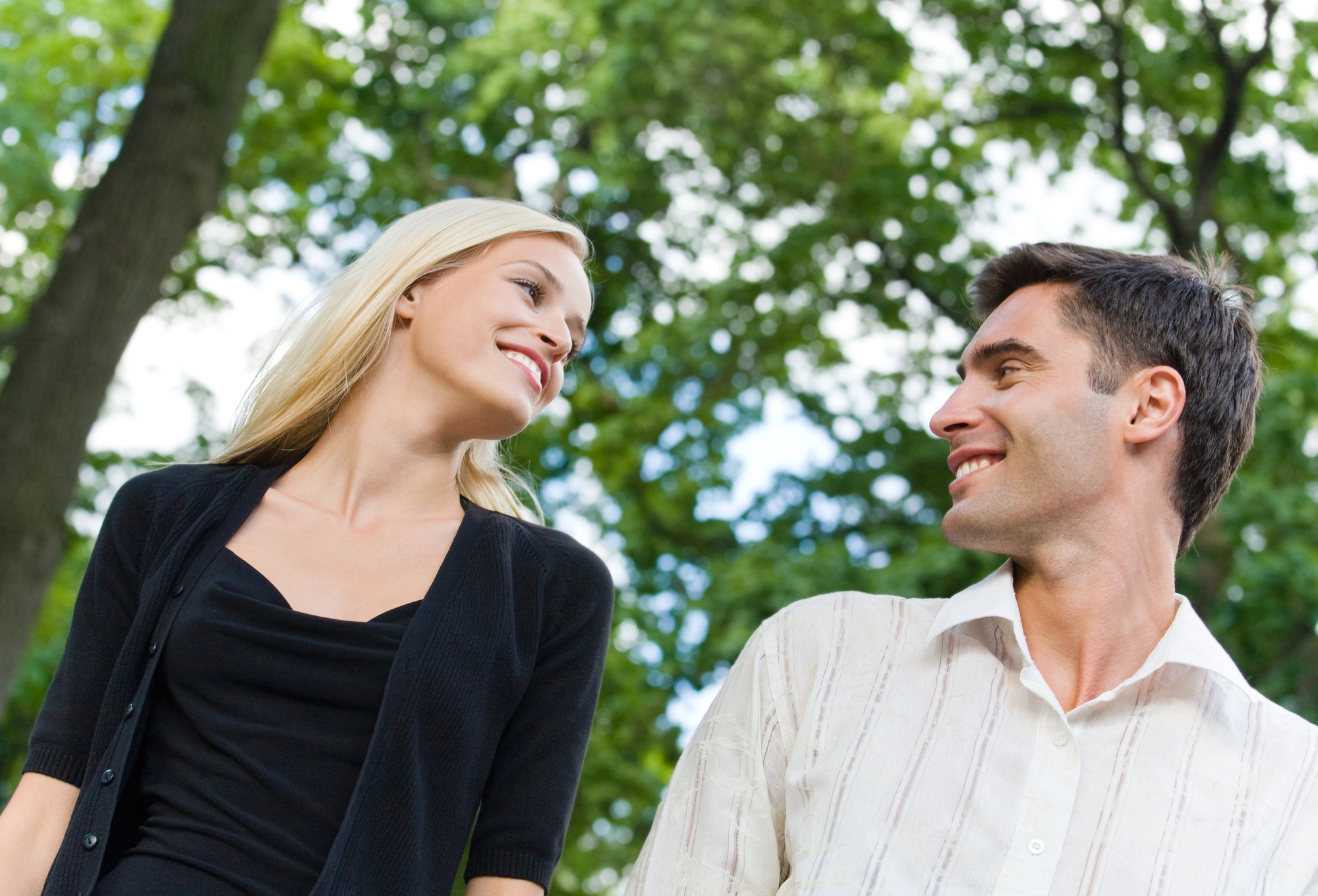 A woman with long blonde hair wearing a black top and cardigan smiles at a man with short dark hair wearing a white striped shirt. They are standing outdoors, surrounded by tall trees with green leaves on a sunny day.