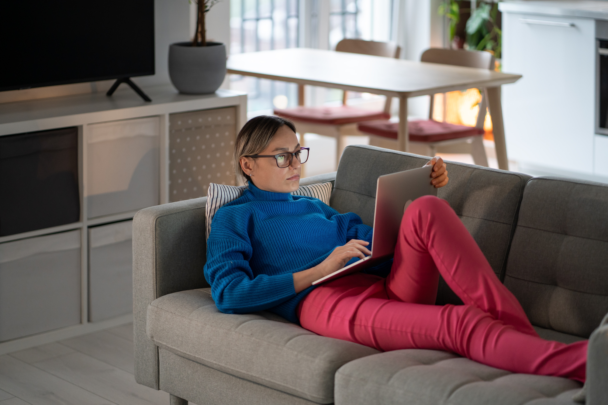 A person wearing glasses, a blue sweater, and red pants is lying on a gray sofa using a laptop. The room features a light-colored interior with a TV, a dining table, and some plants in the background.