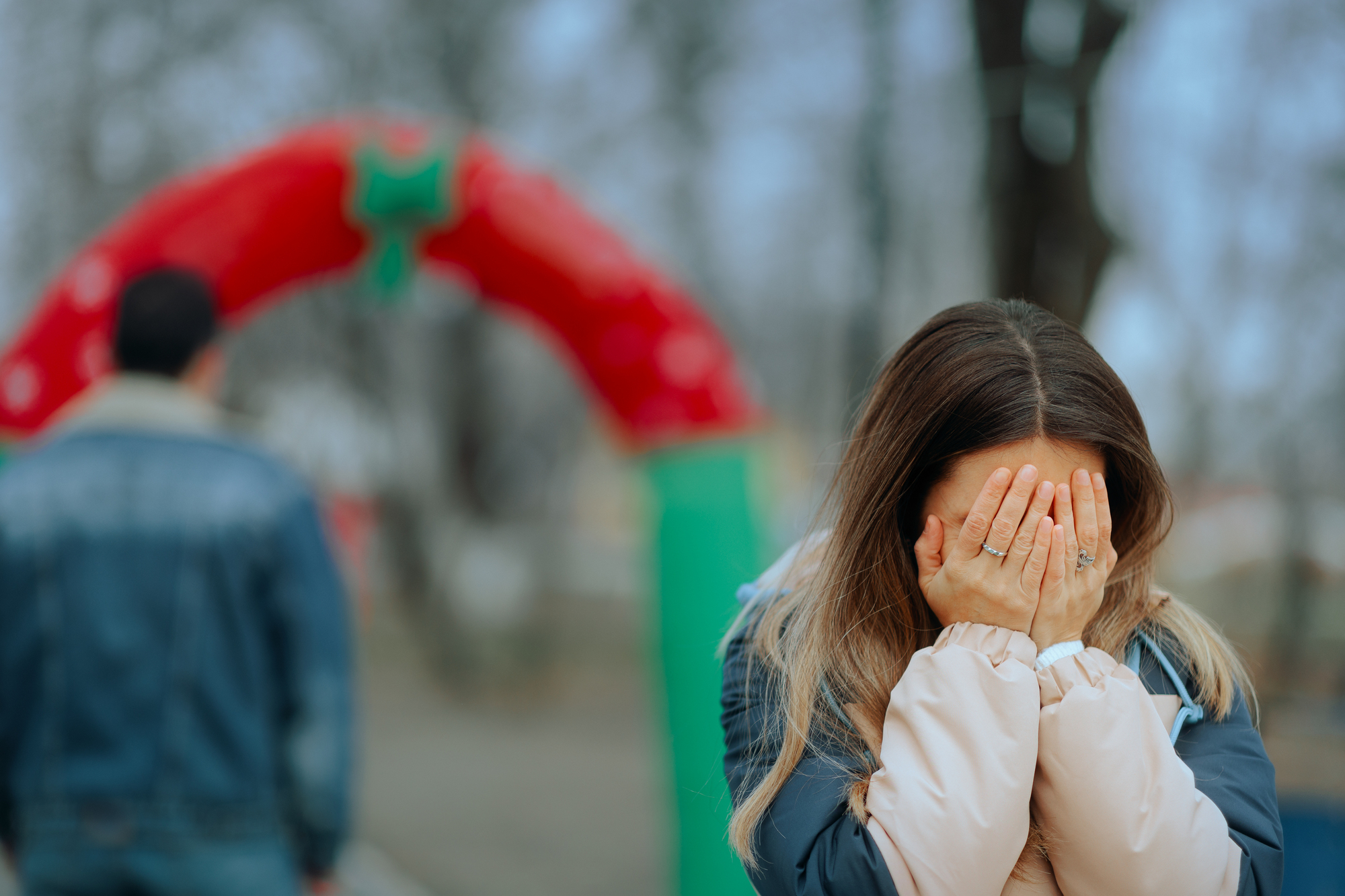 A woman with long hair covers her face with her hands, standing outdoors near a red and green archway. A person in a denim jacket walks away in the background. The scene appears emotional and the weather seems cold.