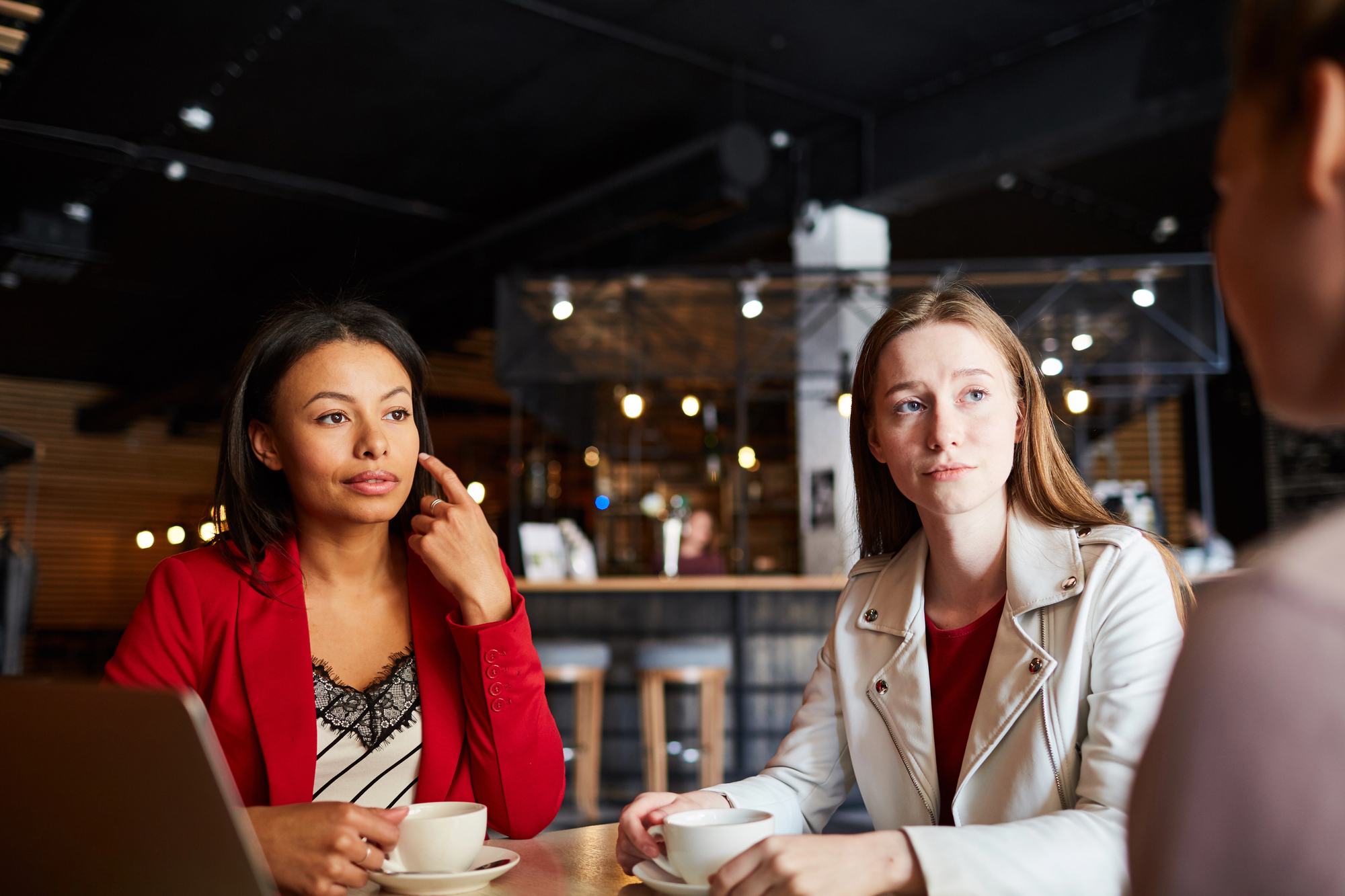 Three women are sitting at a table in a cafe, engaging in conversation. The woman on the left is gesturing with her hand while holding a white cup. They are calmly discussing something, and the background shows a warmly lit cafe environment.