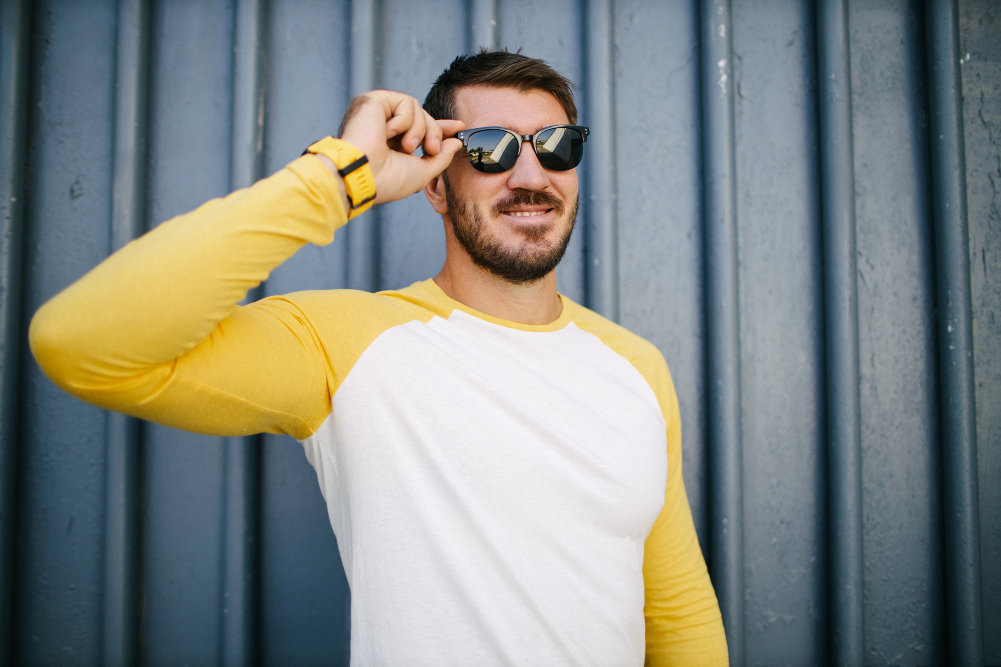 A man with a beard and sunglasses smiles while touching the side of his glasses. He wears a white and yellow long-sleeve shirt and a yellow watch. He stands in front of a vertical corrugated metal wall painted in a grayish-blue color.