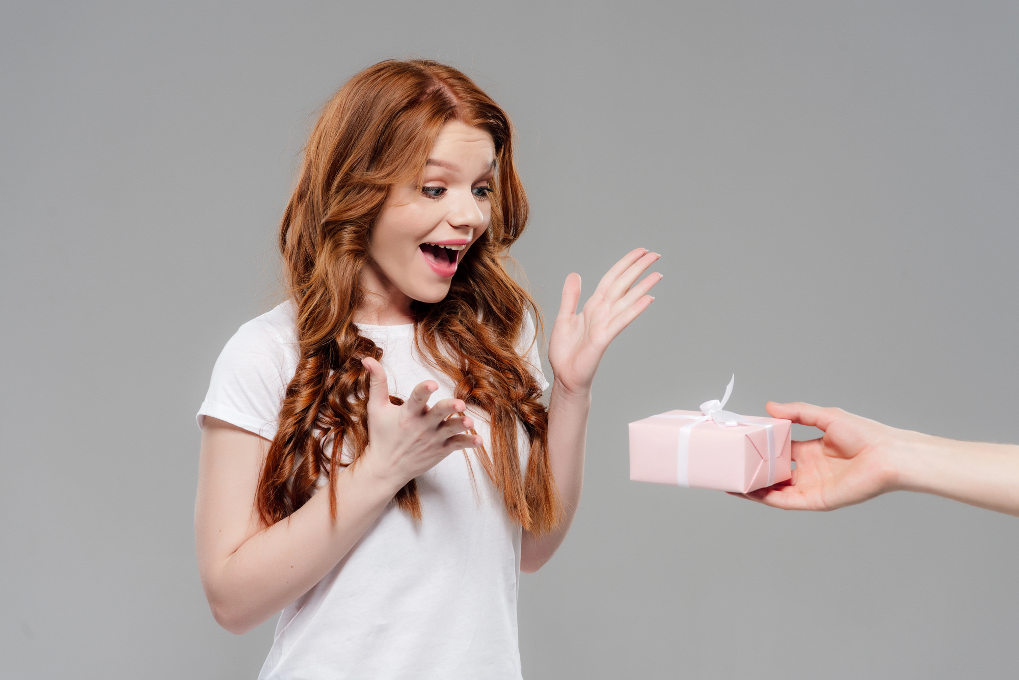 A woman with long red hair and a white shirt looks surprised and delighted as she receives a small pink gift box with a white ribbon from an outstretched hand against a plain gray background.