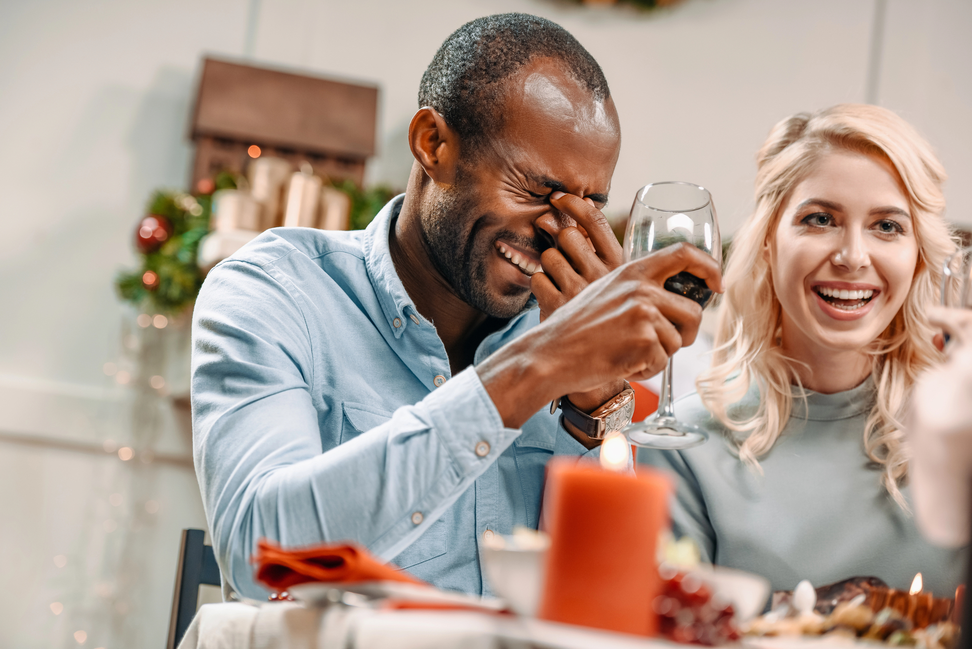 A man and a woman are sitting at a table, enjoying a festive meal. The man is laughing and covering his face with his hand while holding a wine glass. The woman beside him is smiling. The table is decorated with candles, ornaments, and plates of food.