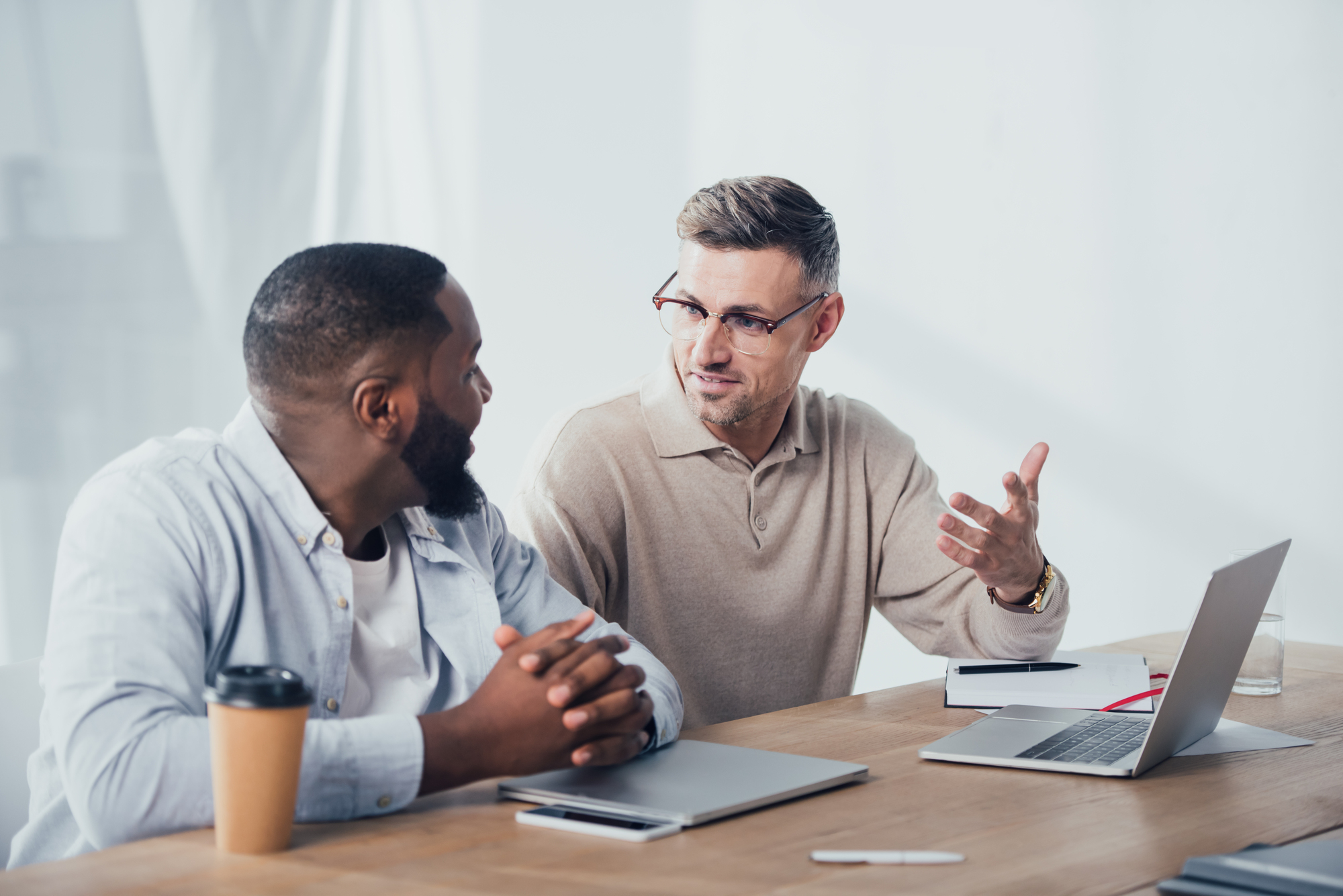 Two men sitting at a desk in a discussion. One man with a beard is wearing a light blue shirt and has a disposable coffee cup in front of him. The other man, with glasses and a gray sweater, gestures with his hand as he speaks. Both have laptops on the desk.