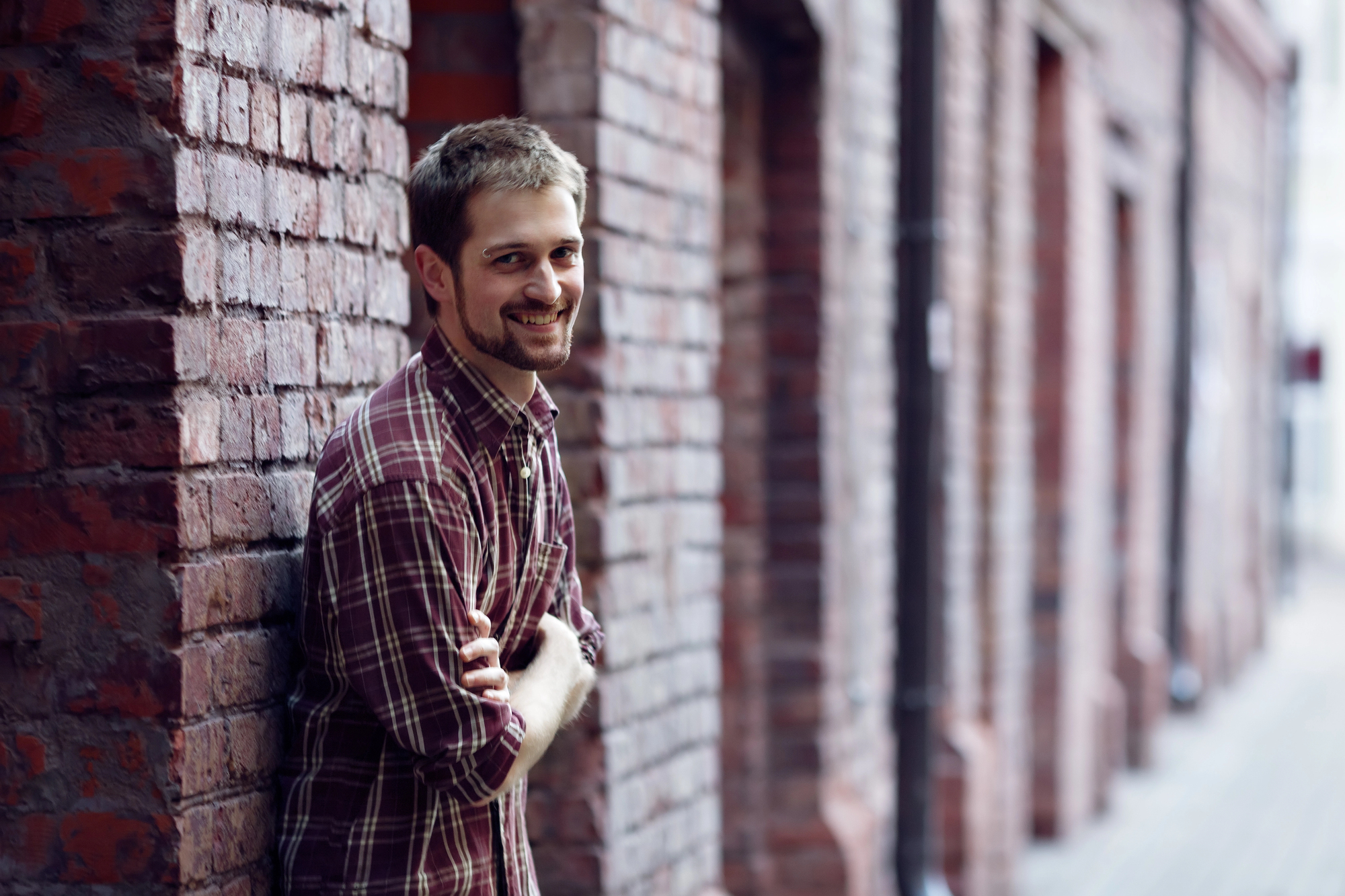 A man with short hair and a beard stands against a brick wall, smiling. He is wearing a plaid shirt and has his arms crossed. The background features a row of brick buildings with windows and doors. The scene is outdoors and bright.