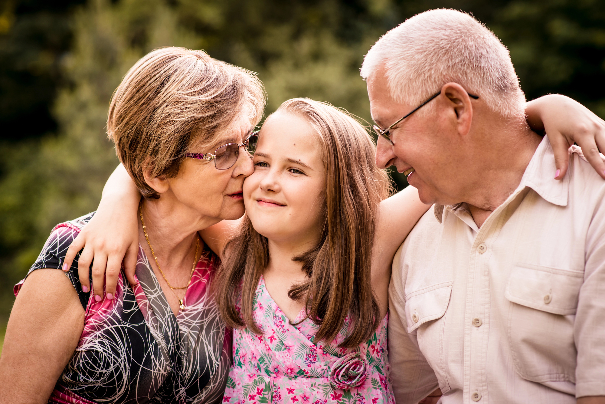 A young girl with shoulder-length brown hair stands between an elderly woman and man, her arms around their shoulders. The woman has short, light brown hair and glasses, while the man has short gray hair and glasses. They all smile warmly at each other against a green, blurry outdoor background.