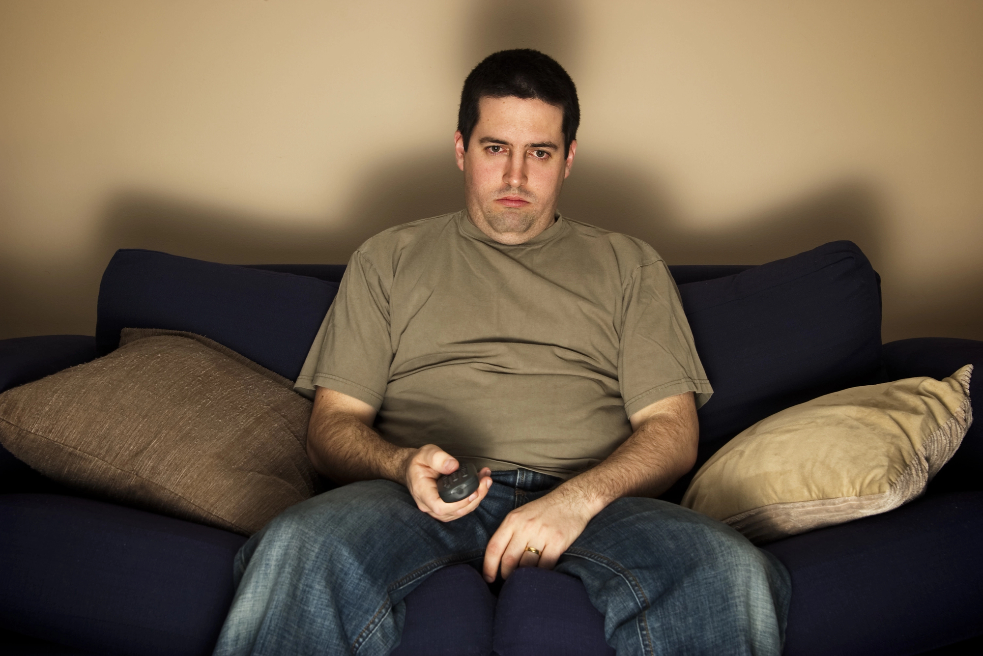 A man wearing a beige t-shirt and jeans is sitting on a dark blue couch with pillows, holding a remote control and staring straight ahead with a neutral or slightly displeased expression. The background is plain beige.