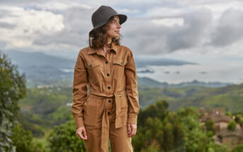 A woman stands outdoors wearing a tan jumpsuit and a dark hat, looking to her right. She is surrounded by a scenic view of rolling green hills, scattered clouds, and distant water bodies.