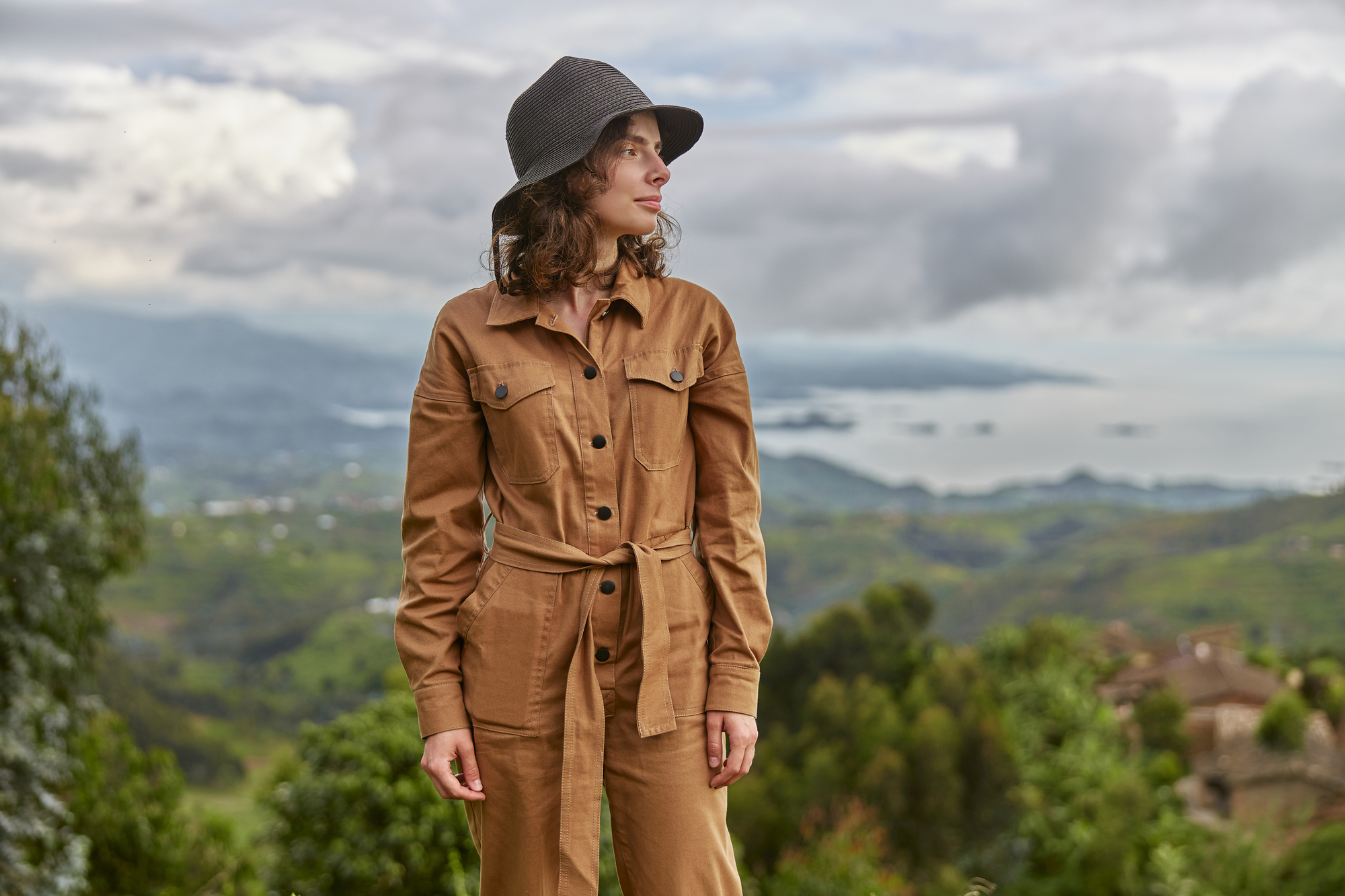 A woman stands outdoors wearing a tan jumpsuit and a dark hat, looking to her right. She is surrounded by a scenic view of rolling green hills, scattered clouds, and distant water bodies.