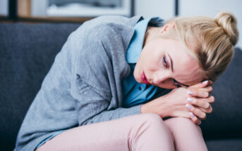 A person with blonde hair is sitting on a couch, leaning forward with their head resting on their clasped hands. They are wearing a light blue shirt and a gray cardigan, and have a thoughtful or worried expression.