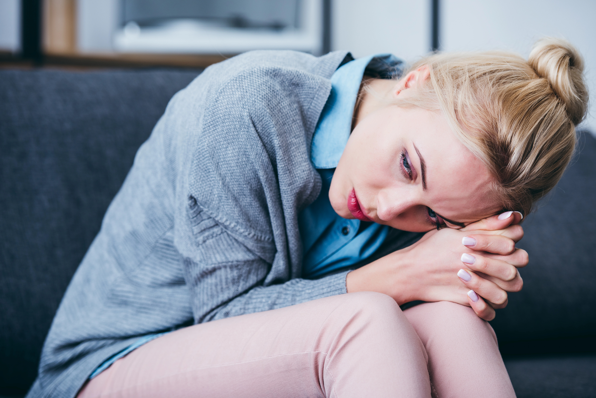 A person with blonde hair is sitting on a couch, leaning forward with their head resting on their clasped hands. They are wearing a light blue shirt and a gray cardigan, and have a thoughtful or worried expression.