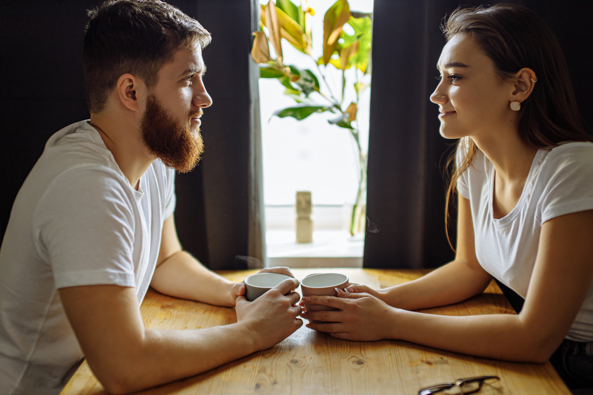 A man and a woman sit across from each other at a wooden table, holding white mugs and smiling. They are both wearing white shirts. A bright window with greenery in the background illuminates the scene. Glasses rest on the table near the woman.