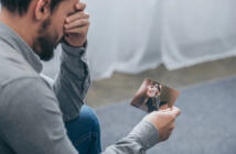 A man with a beard and a grey shirt is sitting and holding a photo of a smiling woman, covering his face with his other hand in an expression of sadness or deep contemplation. The background is blurred and neutral in color.