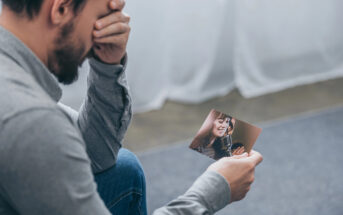 A man with a beard and a grey shirt is sitting and holding a photo of a smiling woman, covering his face with his other hand in an expression of sadness or deep contemplation. The background is blurred and neutral in color.