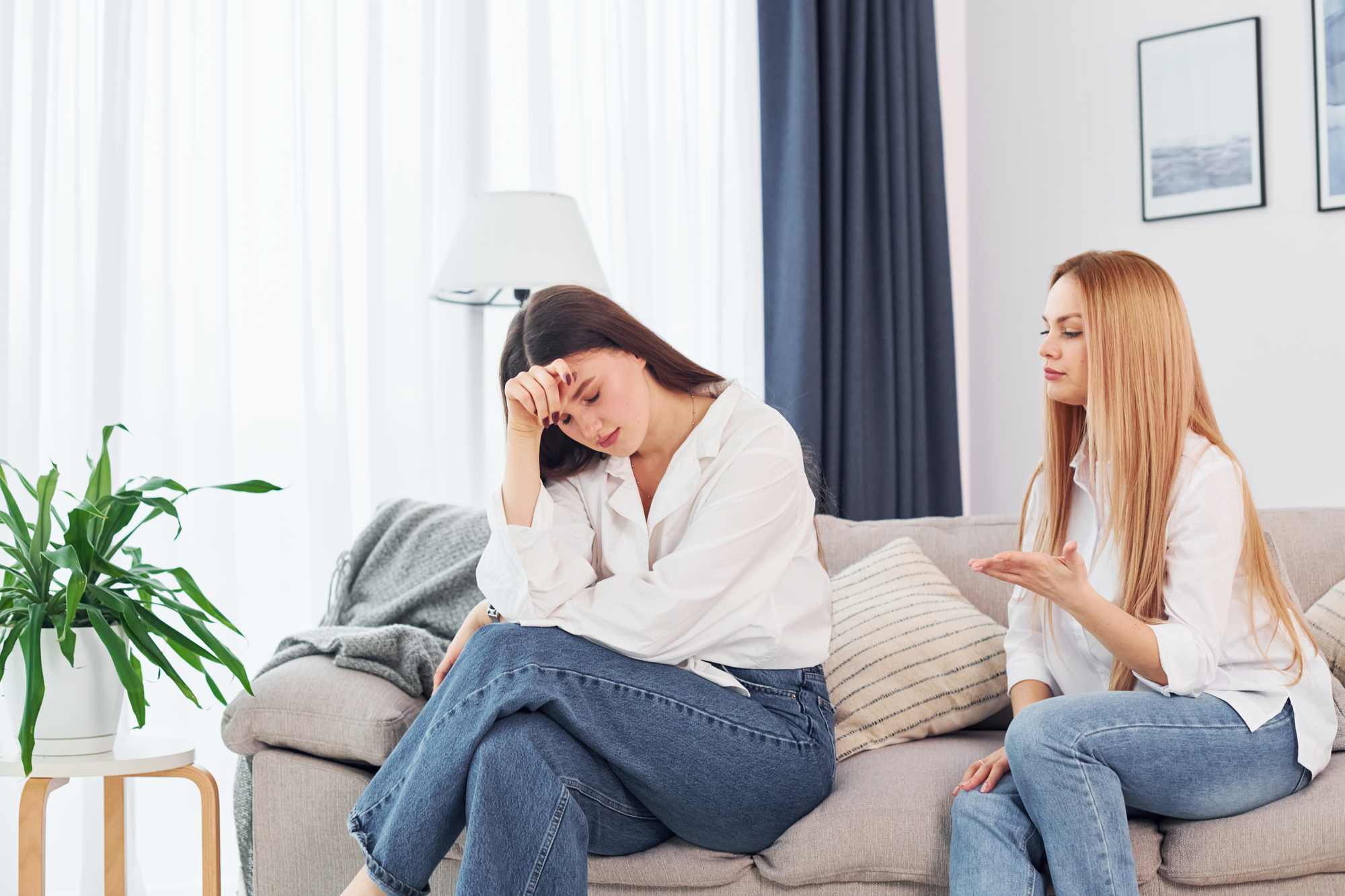 Two women sitting on a couch, engaged in a serious conversation. The woman with long brown hair looks upset, resting her head on her hand, while the woman with long blonde hair appears to be consoling or advising her. The room is bright and minimally decorated.