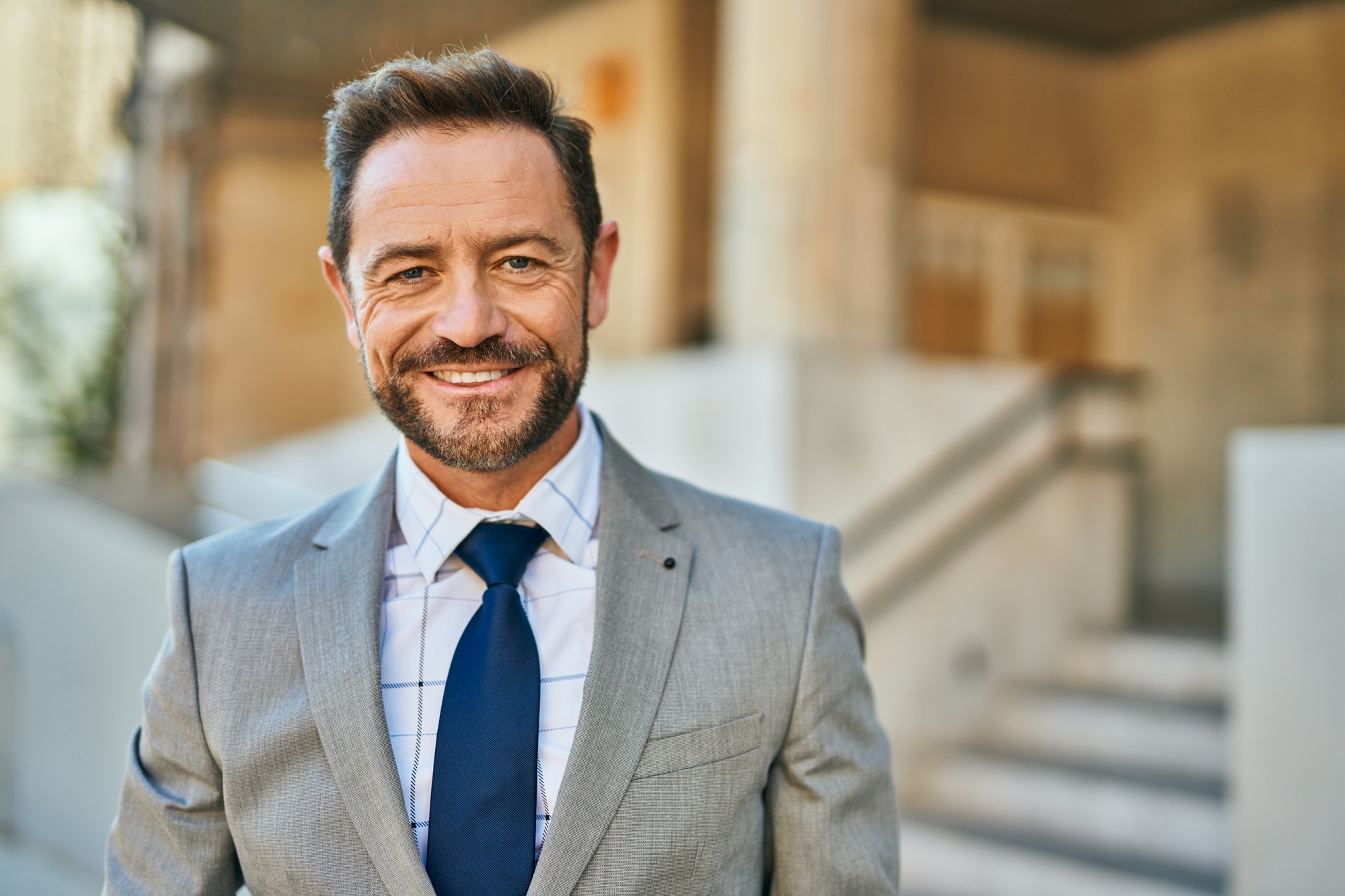 A man in a grey suit, white shirt with a checkered pattern, and blue tie smiles at the camera. He has short brown hair and a beard. The background is an outdoor urban setting with stairs and a blurred building.