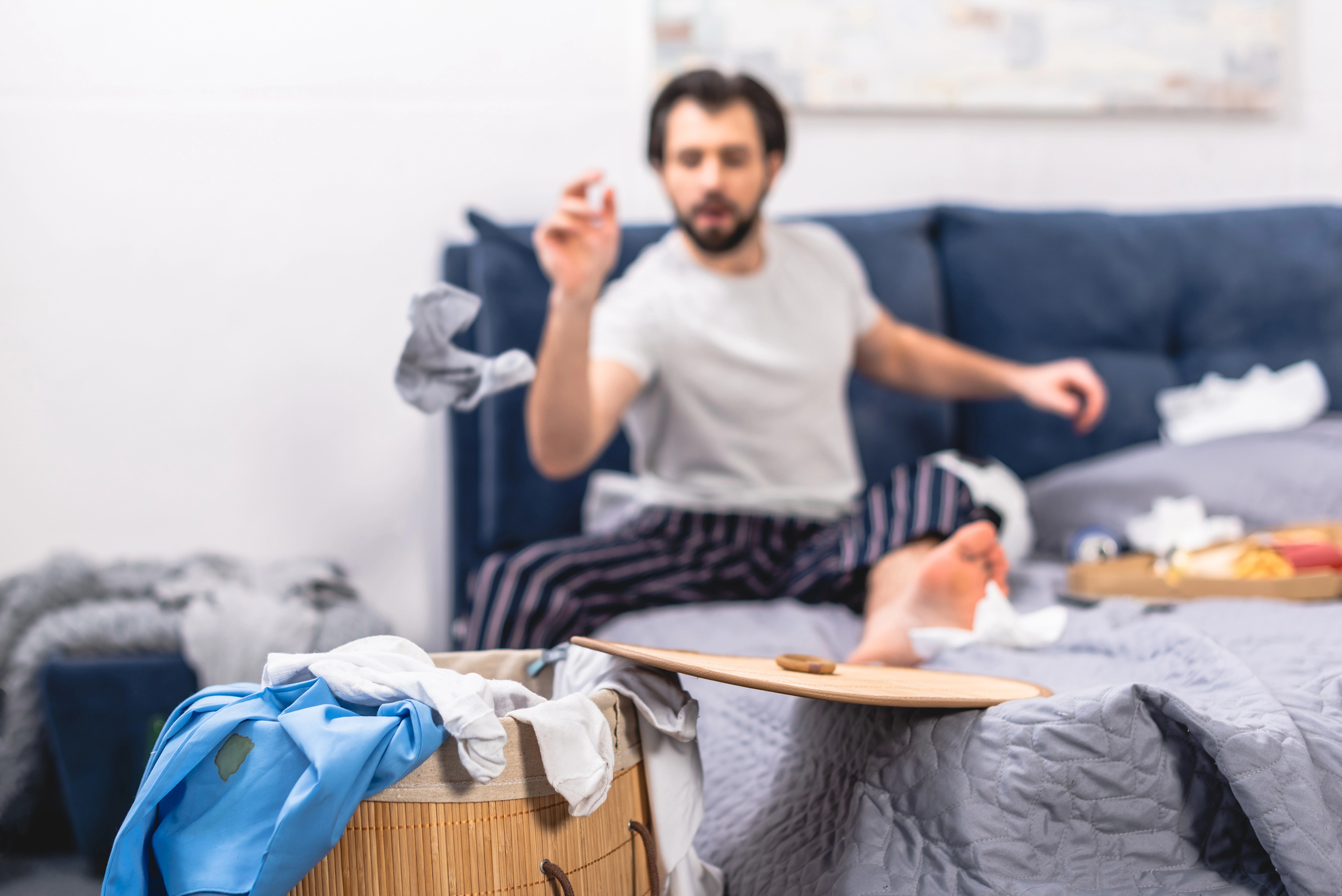 A man in pajamas is sitting on a messy bed, tossing a rolled-up piece of clothing towards an overflowing laundry basket. The background shows a disorganized room with clothes and other items scattered around.