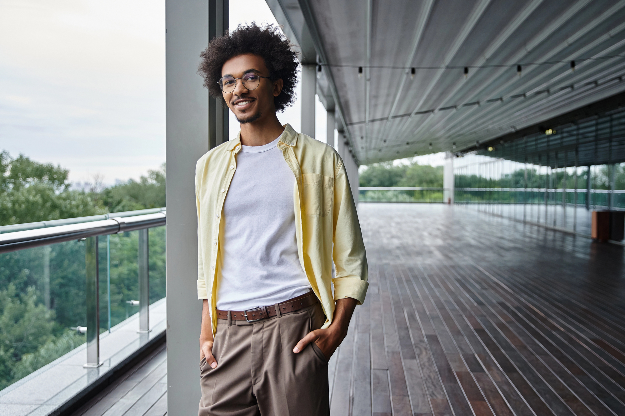 A person with curly hair and glasses, dressed in a light yellow shirt over a white t-shirt and brown pants, stands smiling on a balcony with wooden flooring. The balcony overlooks a green, wooded area and has a metal railing and a roof with hanging lights.